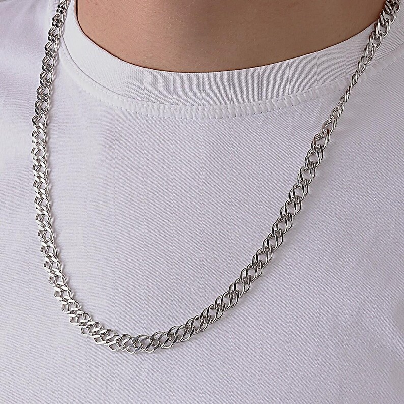 May include: A silver-toned chain necklace featuring a double-link design with a polished, reflective finish. The chain rests against a plain white fabric background, showcasing the intricate, interlocking metal links that form a continuous pattern.