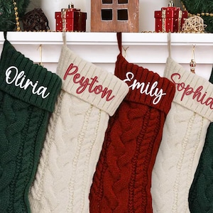 May include: Five cable-knit Christmas stockings in green, cream, and red, each with a name embroidered on the cuff: Olivia, Peyton, Emily, Sophia, and Theodore. The stockings are hanging from a white mantel.