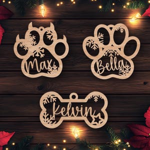 May include: Three wooden Christmas ornaments in the shape of a paw print, a bone, and another paw print. The paw prints have snowflakes and the names "Max" and "Bella" on them. The bone has snowflakes and the name "Kelvin" on it.