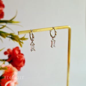 Könnte beinhalten: Silberne Creolen mit rechteckigen, klaren Edelsteinen. Die Ohrringe werden auf einem goldfarbenen Ständer präsentiert, mit roten und grünen floralen Akzenten. Der Markenname "EDELZEIT" ist sichtbar.