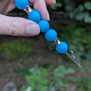 May include: A blue beaded clip with silver-colored accents. The clip has four round, light blue beads connected by silver-toned metal. The clip is held against a blurred green background.