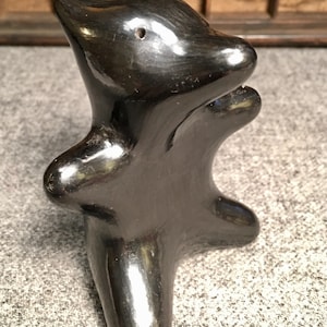 May include: A glossy black ceramic sculpture of an animal-like figure. The sculpture has a rounded body with outstretched arms and a head with a small hole. The piece is displayed on a textured gray surface.