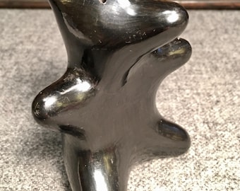 May include: A glossy black ceramic sculpture of an animal-like figure. The sculpture has a rounded body with outstretched arms and a head with a small hole. The piece is displayed on a textured gray surface.