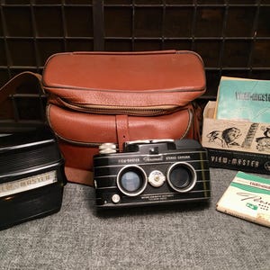 May include: A vintage View-Master stereo camera set, including a black camera, a brown leather case, and View-Master reels. The set also includes a black View-Master viewer and a box of reels. The items are arranged on a gray surface.