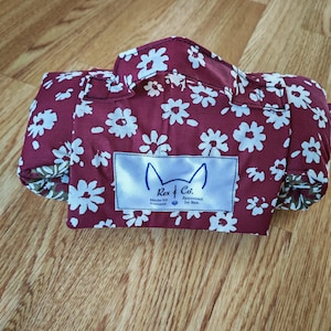 May include: A rolled fabric item featuring a deep burgundy background with a white floral pattern. It includes a top handle and a rectangular white label with the text Rex & Co. Made by humans. Approved by Rex, featuring a cat ear icon.