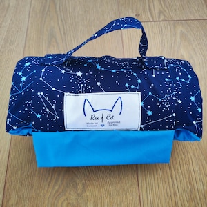 May include: A rolled fabric carrier featuring a dark blue celestial print with white stars and constellations. It includes a solid bright blue bottom panel, a matching handle, and a white rectangular label reading Rex & Co. Made by humans. Approved by Rex.