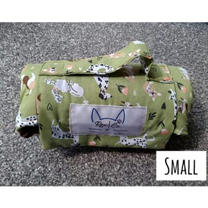 May include: A rolled olive green fabric mat featuring a repeating pattern of various dog breeds, small flowers, and hearts. A rectangular label on the front displays the text Rex & Co. Made by humans. Approved by Rex. The word SMALL is printed in black.