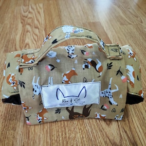 May include: A tan fabric bag with a handle featuring a repeating pattern of various dog breeds, small flowers, and hearts. A white rectangular label on the front displays the text Rex & Co. Approved by Rex with a line drawing of dog ears.