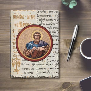 May include: A hardcover journal with a religious theme. The cover features an illustration of a bearded man writing, surrounded by a red and gold circular frame. The background is filled with handwritten text in a vintage style.