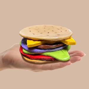 May include: A vibrant, handmade burger featuring layers of red, green, purple, yellow, and brown ingredients, topped with a tan bun. The burger is held in a hand against a neutral backdrop. The burger is a playful, artistic depiction of a classic food item.