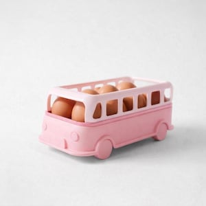 May include: A pink plastic egg holder in the shape of a bus, containing brown eggs. The bus features a rectangular top with open windows and circular wheels. The eggs are visible through the windows.