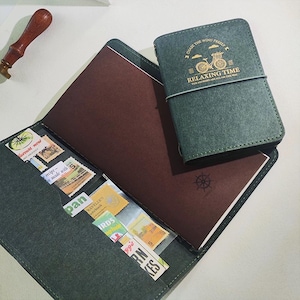 Könnte beinhalten: Ein teal-farbenes Notizbuch mit dem Text "CHASE THE WIND FREELY" und "RELAXING TIME" auf dem Einband. Das Notizbuch ist geöffnet und zeigt braune Seiten und eine Tasche voller bunter Briefmarken. Ein Holzstempel ist sichtbar.