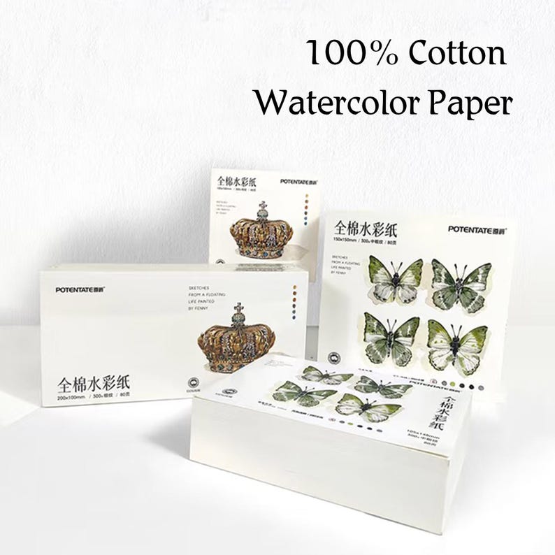 K&ouml;nnte beinhalten: Wei&szlig;e Aquarellpapierboxen und -bl&ouml;cke mit Schmetterlings- und Kronenillustrationen. Der Text auf den Boxen lautet "100% Cotton Watercolor Paper" und "POTENTATER". Die Bl&ouml;cke zeigen Schmetterlings- und Kronenmotive.