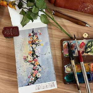 May include: A watercolor painting of a floral arrangement with pink, orange, and yellow flowers on a light blue background. The painting is on a small notepad. A wooden palette with paint and paintbrushes are also visible.