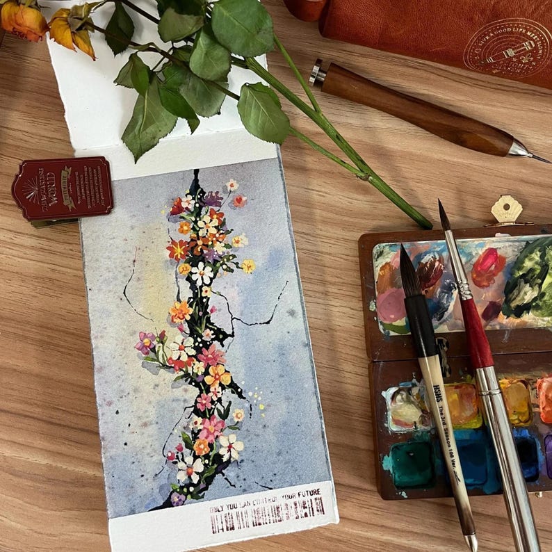 May include: A watercolor painting of a floral arrangement with pink, orange, and yellow flowers on a light blue background. The painting is on a small notepad. A wooden palette with paint and paintbrushes are also visible.