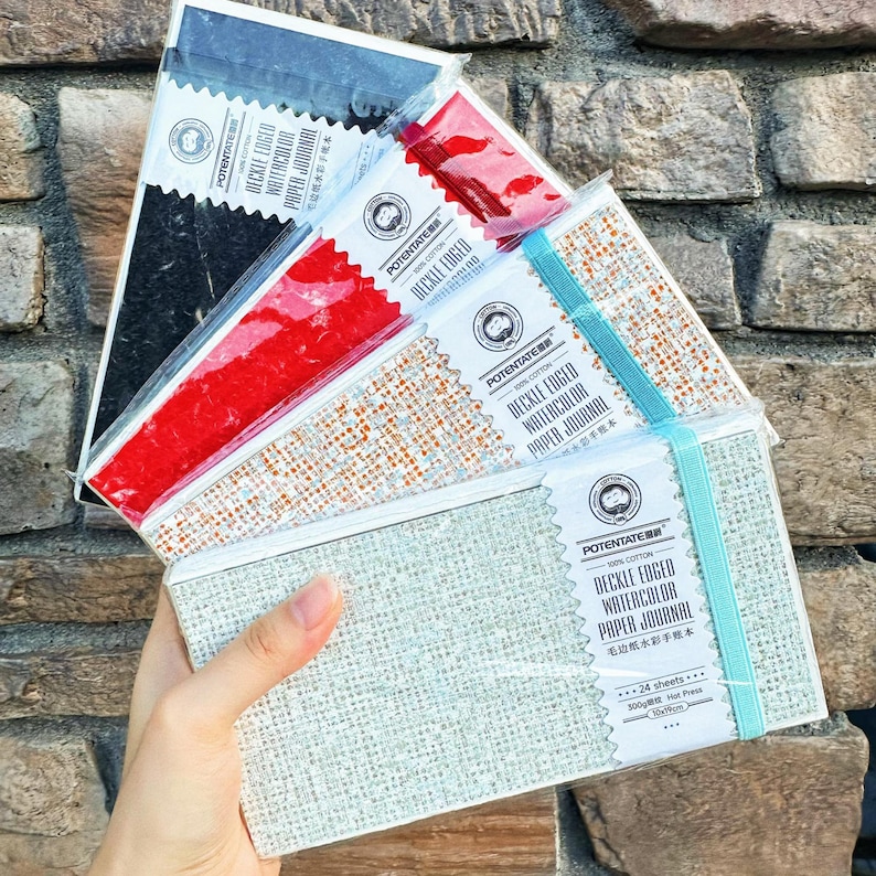 May include: A hand holds a stack of watercolor paper journals. The journals have deckle edges and are bound with elastic bands. The covers are various colors, including gray, red, and black. Each journal has the text "POTENTATE DECKLE EDGED WATERCOLOR PAPER JOURNAL".