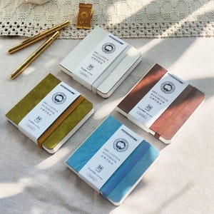 Op de afbeelding: Vier notitieboeken met verschillende gekleurde covers: wit, groen, bruin en blauw. Elk notitieboek heeft een bijpassende elastische band en een wit label met tekst. De notitieboeken zijn gelabeld met "100% Cotton Handbook".