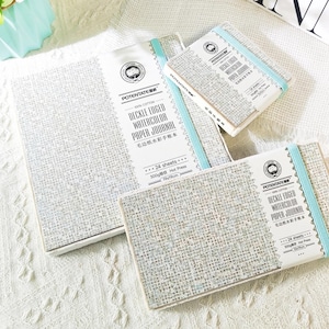 May include: Several watercolor paper journals are displayed, varying in size. The journals have a textured, light gray cover with a white label that reads "DECKLE EDGED WATERCOLOR PAPER JOURNAL." Each journal is bound with a light blue elastic band. The text on the label includes "100% Cotton" and "24 sheets."