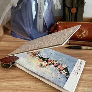 May include: A watercolor journal with a floral design, held open with a metal clip. The journal's cover features a textured pattern. Other items include a leather pouch, a pen, and a wrapped gift.
