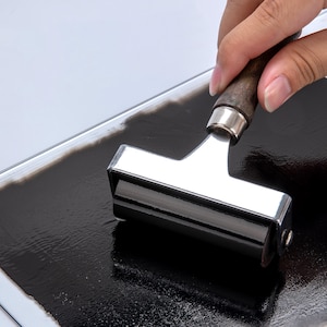 Puede incluir: Primer plano de un rodillo de tinta negro que se utiliza para extender tinta negra sobre una superficie de vidrio. El rodillo está unido a un mango de metal.