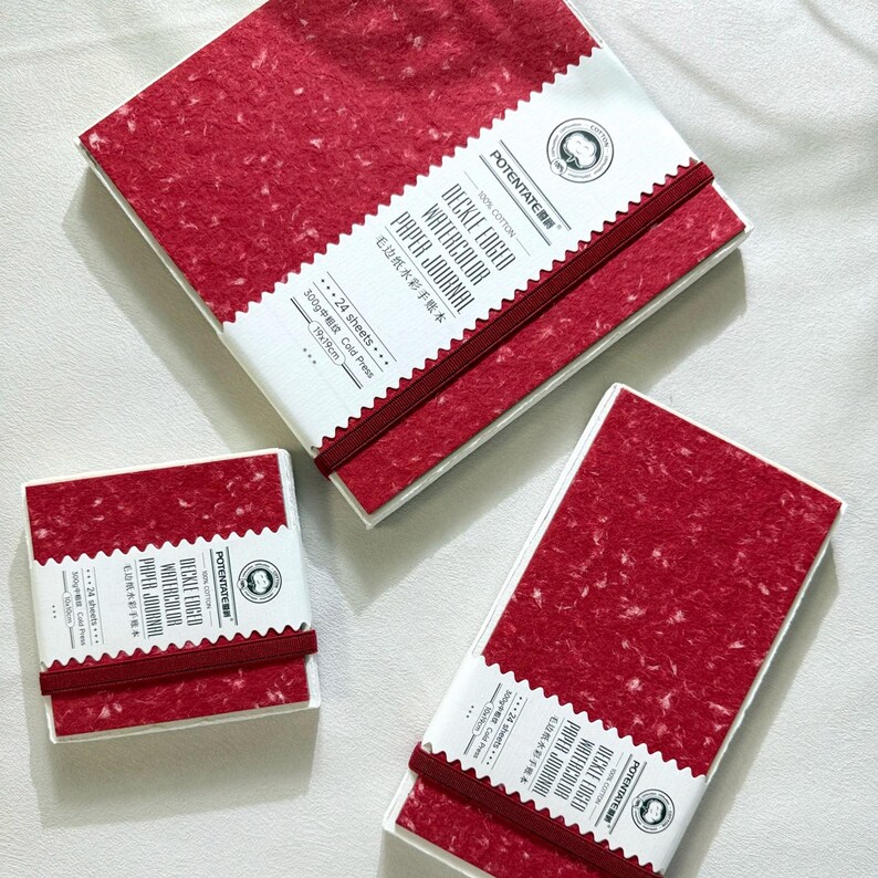 May include: Three watercolor paper journals with red speckled covers and white labels. The labels read "DECKLE EDGED WATERCOLOR PAPER JOURNAL" and include the brand name "POTENTATE". Each journal is bound with a red elastic band. The journals are 19cm x 19cm.