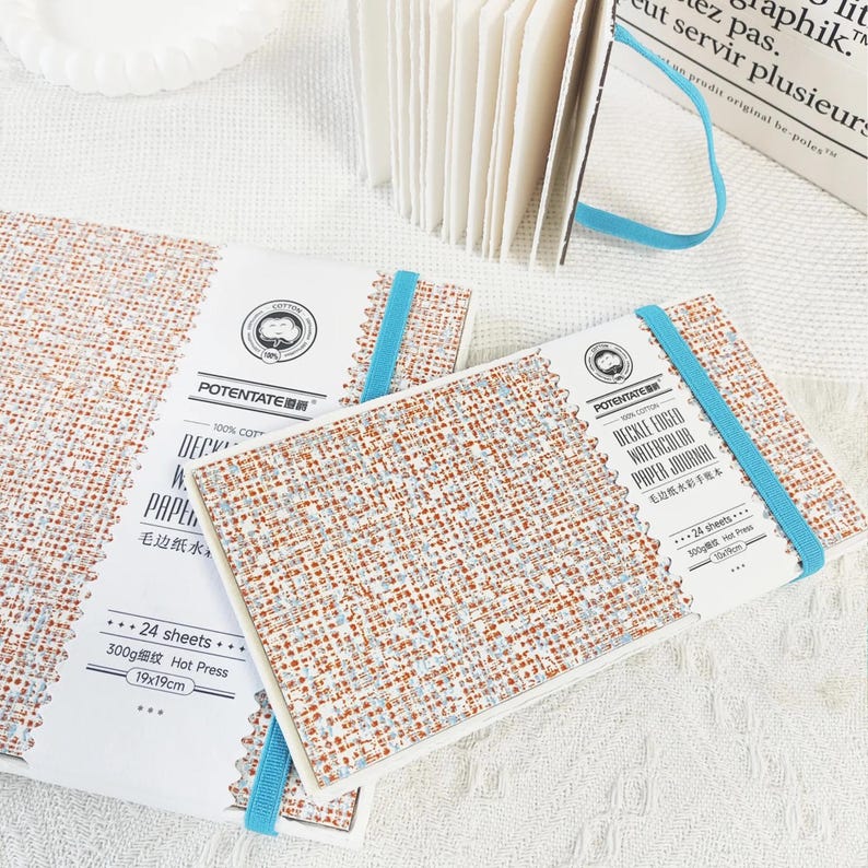May include: Watercolor paper journals with a textured, woven-like cover in shades of brown, white, and blue. Each journal is secured with a light blue elastic band. The cover features the text "POTENTATE" and "DECKLE EDGED WATERCOLOR PAPER JOURNAL". The journals measure 19x19cm.