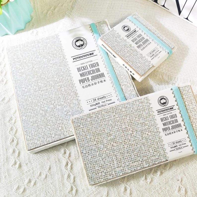 May include: A collection of watercolor paper journals in various sizes. The journals are light gray with a textured surface and are bound with a light blue elastic band. Each journal has a white label with text that reads "DECKLE EDGED WATERCOLOR PAPER JOURNAL".