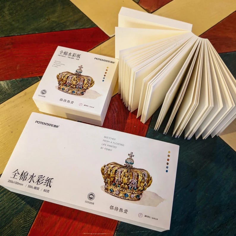 K&ouml;nnte beinhalten: Wei&szlig;e Aquarellpapierbl&ouml;cke mit einer goldenen Kronenillustration und chinesischem Text. Ein Block ist ge&ouml;ffnet und zeigt die einzelnen Bl&auml;tter. Die Bl&ouml;cke liegen auf einer mehrfarbigen Holzoberfl&auml;che.