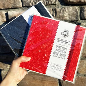 May include: Two watercolor paper journals. One journal has a red cover with white text and a red ribbon. The other journal has a black cover. The text on the red journal reads "Deckle Edged Watercolor Paper Journal" and "19x19cm".