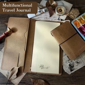 Op de afbeelding: Open bruin leren reisdagboek met de tekst "Multifunctioneel Reisdagboek". Het dagboek is open en toont lege pagina's en een kleine vliegtuigillustratie. Een kleiner dagboek is gesloten met een zwarte elastische band. Andere items zijn een kompas en aquarelverf.