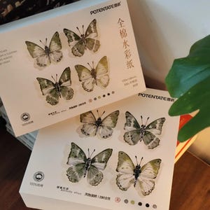 K&ouml;nnte beinhalten: Zwei Stapel wei&szlig;es Papier mit Aquarell-Schmetterlingsdesigns. Jedes Blatt zeigt sechs Schmetterlinge in Gr&uuml;n-, Braun- und Wei&szlig;t&ouml;nen. Text in chinesischen Schriftzeichen und Englisch ist auf der Verpackung zu sehen.