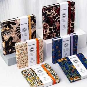 May include: A selection of notebooks with various floral and botanical designs. The notebooks display patterns such as roses, leaves, and citrus fruits. Each notebook is bound with an elastic band and a white paper band with the text "100% Cotton Handbook".