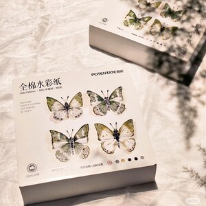 K&ouml;nnte beinhalten: Eine wei&szlig;e Schachtel Aquarellpapier mit Schmetterlingsillustrationen. Die Schachtel tr&auml;gt den Text "POTENTATE" und chinesische Schriftzeichen. Die Schmetterlinge sind in Gr&uuml;n, Wei&szlig; und Braun gehalten. Das Papier misst 150x150mm.