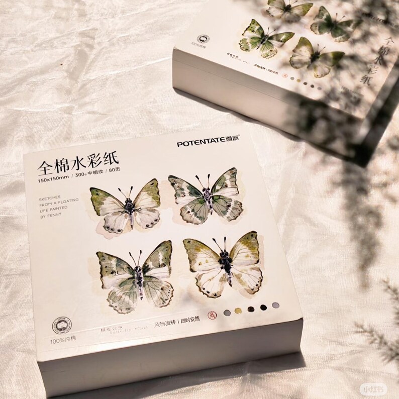 K&ouml;nnte beinhalten: Eine wei&szlig;e Schachtel Aquarellpapier mit Schmetterlingsillustrationen. Die Schachtel tr&auml;gt den Text "POTENTATE" und chinesische Schriftzeichen. Die Schmetterlinge sind in Gr&uuml;n, Wei&szlig; und Braun gehalten. Das Papier misst 150x150mm.