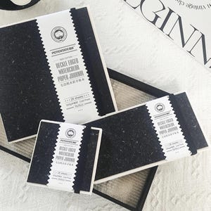 May include: Several black watercolor paper journals with white labels. The labels read "DECKLE EDGED WATERCOLOR PAPER JOURNAL" and include the text "100% COTTON". Each journal has 24 sheets and is 19x19cm.