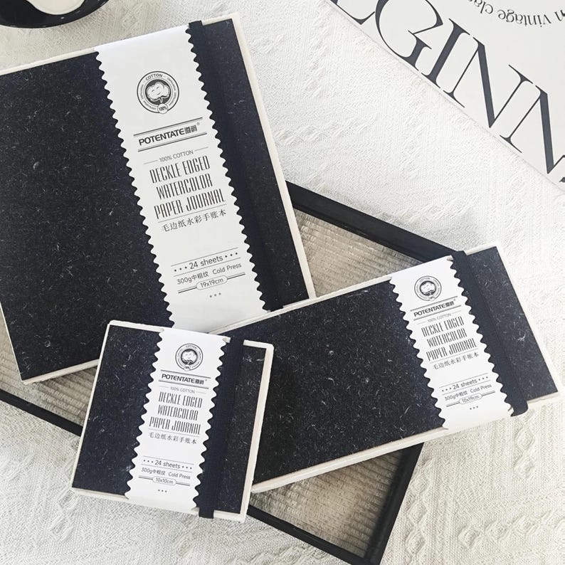 May include: Several black watercolor paper journals with white labels. The labels read "DECKLE EDGED WATERCOLOR PAPER JOURNAL" and include the text "100% COTTON". Each journal has 24 sheets and is 19x19cm.