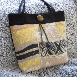 Puede incluir: Un bolso tote negro con un diseño de patchwork de tela beige, amarilla y negra. El bolso tiene una correa de cuero negra y un cierre de botón marrón.