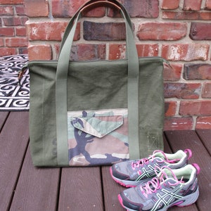 May include: A green canvas tote bag with a camouflage pocket and leather handles. Two gray running shoes with pink laces are in front of the bag.