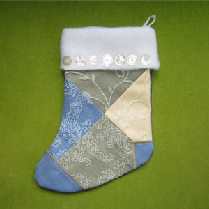 May include: A patchwork Christmas stocking featuring a white faux fur cuff adorned with seven clear buttons. The body is constructed from triangular fabric panels in shades of sage green, light blue, and cream, each featuring intricate floral embroidery.