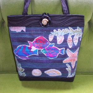 May include: A black tote bag with a dark blue and green fabric depicting a sea life scene with colorful fish, seashells, and a starfish. The bag has a black fabric handle and a decorative button.