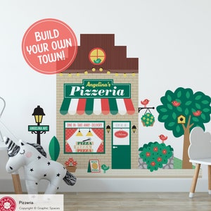 May include: A wall decal set featuring a brick-patterned pizzeria building with a red, white, and green striped awning, a sign reading Angelina's Pizzeria, a street lamp labeled Angelina Ave, a green tree, and small red birds and flowers.