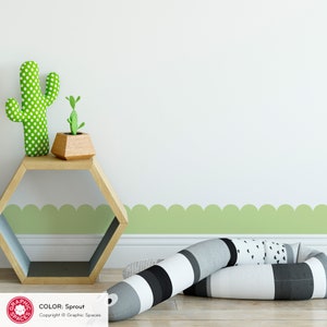 May include: A green and white polka dot cactus and a small green plant in a wooden pot sit on a hexagonal wooden shelf. A green scalloped border runs along the bottom of a white wall. A black and white striped pillow sits on the floor.