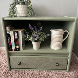 Könnte beinhalten: Ein grünes Holzregal mit einer Schublade. Das Regal hat eine weiße Kanne, eine Topfpflanze mit lila Blüten und eine Topfpflanze mit grünen Blättern. Die Schublade hat ein Blumenmuster und einen silbernen Knopf. Das Regal steht auf einem rosa Teppich.