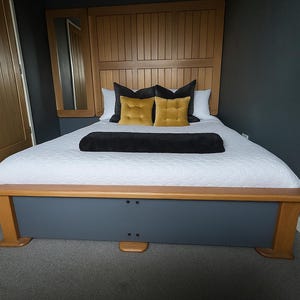 May include: A wooden bed frame with a grey base and light wood trim. The headboard features vertical wooden panels and a side mirror. The bed is made with white bedding, black and yellow pillows, and a black throw. The bed measures approximately 200cm in length.
