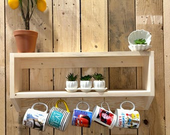 May include: A wooden shelf displays a potted lemon tree, small succulents, and five hanging mugs. The mugs feature various designs, including cartoon characters and the text "A CUP OF TEA!" The shelf is mounted on a wooden plank wall.