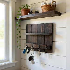 May include: A dark stained wooden key holder with a small shelf and four hooks, mounted on a white wall. A second shelf above holds a potted plant, sunglasses, and a woven basket. Keys hang from the hooks.