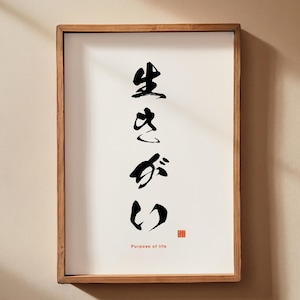 May include: A framed print with Japanese calligraphy in black ink on a white background. The wooden frame surrounds the artwork, which features the word "Ikigai" and the English translation "Purpose of life". The print is displayed on a beige wall.
