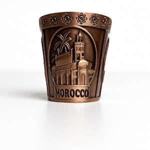 Könnte beinhalten: Bronzefarbenes Schnapsglas mit einem detaillierten Relief einer marokkanischen Stadtlandschaft, einschließlich einer Moschee und Palmen. Das Wort "MOROCCO" ist am Boden abgebildet. Der Rand hat ein sich wiederholendes Blumenmuster.