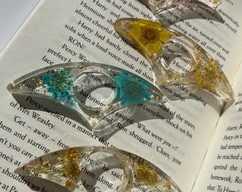May include: Set of three clear resin hair clips with dried flowers and gold flakes. The flowers are purple, yellow, and pink. The clips are shaped like a crescent moon.