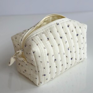 Puede incluir: Una bolsa de maquillaje acolchada blanca con cierre de cremallera y un pequeño estampado floral. La bolsa está hecha de una tela suave y texturizada.
